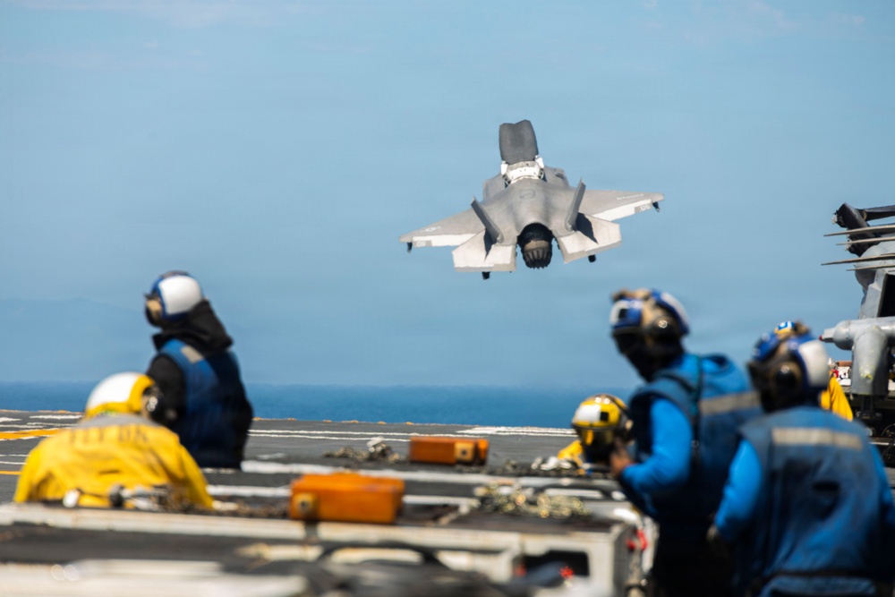 A military jet aircraft, consistent with an F-35B, is performing a short takeoff or vertical landing maneuver above the flight deck of a naval vessel, likely an amphibious assault ship. Multiple flight deck crew members in color-coded jerseys and helmets are observing the aircraft from the deck. The image captures a dynamic carrier aviation operation over open ocean.