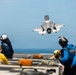 A military jet aircraft, consistent with an F-35B, is performing a short takeoff or vertical landing maneuver above the flight deck of a naval vessel, likely an amphibious assault ship. Multiple flight deck crew members in color-coded jerseys and helmets are observing the aircraft from the deck. The image captures a dynamic carrier aviation operation over open ocean.