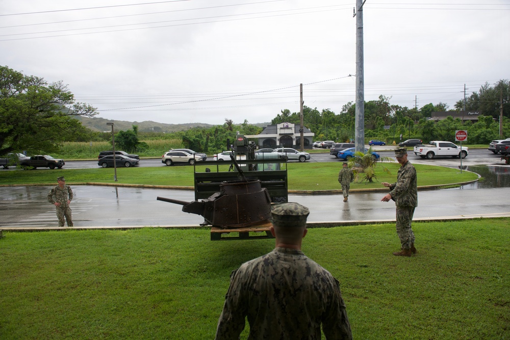 Several military personnel in camouflage uniforms are gathered around what appears to be a historic artillery piece or gun mount displayed on a grass area near a roundabout. The setting appears to be a military base or installation, likely in a tropical location given the vegetation, and the scene is taking place during overcast, rainy conditions. Commercial establishments including what appears to be a Pizza Hut are visible in the background across the road.