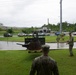 Several military personnel in camouflage uniforms are gathered around what appears to be a historic artillery piece or gun mount displayed on a grass area near a roundabout. The setting appears to be a military base or installation, likely in a tropical location given the vegetation, and the scene is taking place during overcast, rainy conditions. Commercial establishments including what appears to be a Pizza Hut are visible in the background across the road.