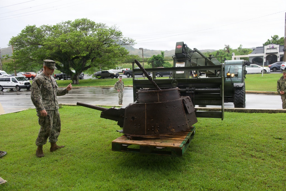 Military personnel in camouflage uniforms are examining a heavily corroded and rusted tank turret placed on a wooden pallet on a grass lawn. The turret appears to be a recovered historical artifact, likely from World War II, being transported or displayed at what appears to be a military installation. A green military forklift/vehicle is visible in the background, suggesting the turret was recently moved.