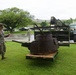 Military personnel in camouflage uniforms are examining a heavily corroded and rusted tank turret placed on a wooden pallet on a grass lawn. The turret appears to be a recovered historical artifact, likely from World War II, being transported or displayed at what appears to be a military installation. A green military forklift/vehicle is visible in the background, suggesting the turret was recently moved.