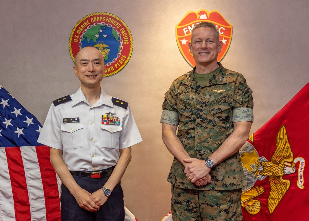 Two military officers pose for a formal photograph in front of institutional insignia. One officer wears a white dress uniform consistent with the Japan Maritime Self-Defense Force, while the other wears U.S. Marine Corps MARPAT camouflage utilities. The setting appears to be an official meeting room at a U.S. Marine Corps Forces Pacific facility.