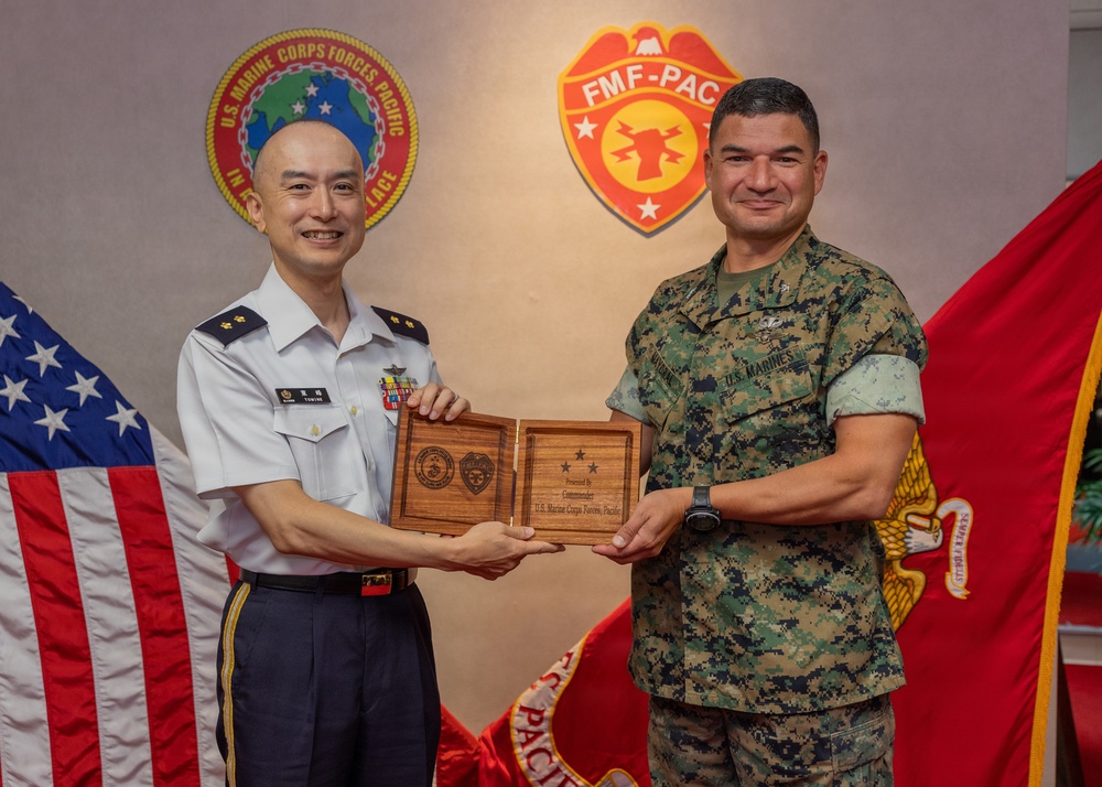 Two military officers are posing together while jointly holding a wooden commemorative plaque. The setting features U.S. Marine Corps Forces Pacific insignia and logos on the wall behind them, along with an American flag and a Marine Corps flag. The officer on the left wears a white dress uniform with what appears to be an Asian military service uniform, while the officer on the right wears U.S. Marine Corps MARPAT camouflage utilities.