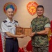 Two military officers are posing together while jointly holding a wooden commemorative plaque. The setting features U.S. Marine Corps Forces Pacific insignia and logos on the wall behind them, along with an American flag and a Marine Corps flag. The officer on the left wears a white dress uniform with what appears to be an Asian military service uniform, while the officer on the right wears U.S. Marine Corps MARPAT camouflage utilities.