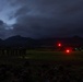 A low-light military field operation captured at dusk or dawn, showing a group of uniformed personnel standing in formation on an open grassy field. A helicopter with active red and green navigation lights is visible in the mid-ground, suggesting a landing zone or staging area. A mountainous terrain with heavy cloud cover forms the backdrop.