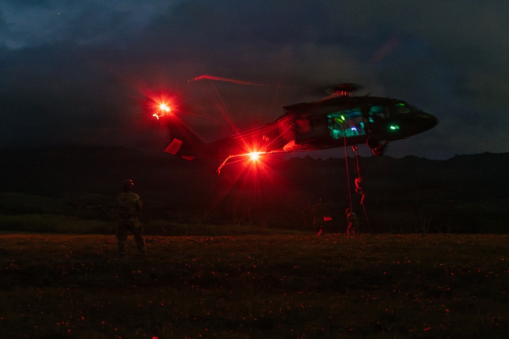A military helicopter, likely a UH-60 Black Hawk, is conducting a nighttime fast-rope or rappelling operation in an open field. Personnel can be seen descending from the aircraft via ropes while at least one soldier stands on the ground observing. The scene is illuminated primarily by the helicopter's red navigation/warning lights, creating a dramatic low-light environment.