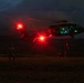 A military helicopter, likely a UH-60 Black Hawk, is conducting a nighttime fast-rope or rappelling operation in an open field. Personnel can be seen descending from the aircraft via ropes while at least one soldier stands on the ground observing. The scene is illuminated primarily by the helicopter's red navigation/warning lights, creating a dramatic low-light environment.