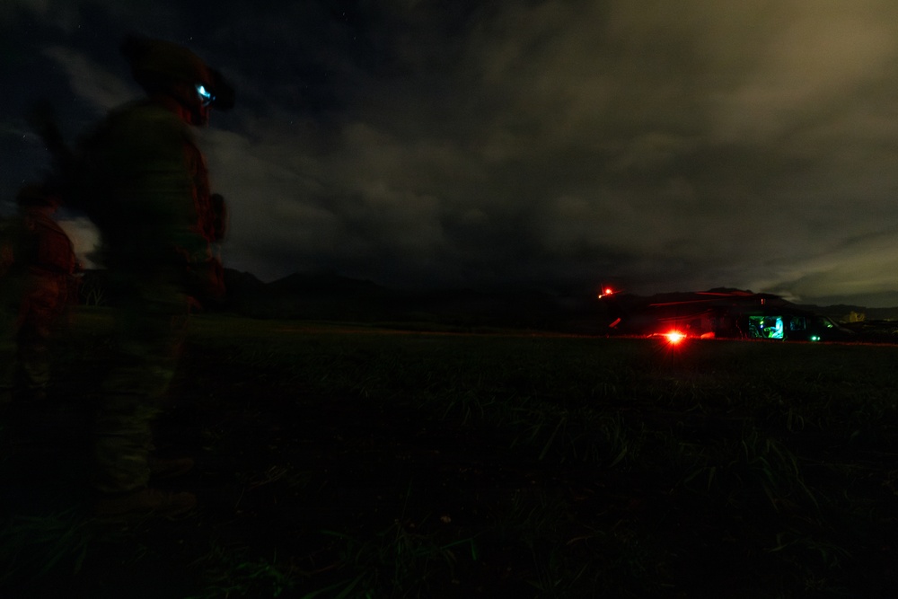 A nighttime military operation scene showing a soldier in full gear on the left side with an illuminated helmet-mounted device, observing a helicopter on a landing zone in the distance. The helicopter displays standard aviation navigation lights including red and green position lights. The scene is set on an open grassy field with mountainous terrain visible in the background under a cloudy night sky.