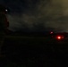A nighttime military operation scene showing a soldier in full gear on the left side with an illuminated helmet-mounted device, observing a helicopter on a landing zone in the distance. The helicopter displays standard aviation navigation lights including red and green position lights. The scene is set on an open grassy field with mountainous terrain visible in the background under a cloudy night sky.