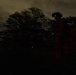 A nighttime military operation scene showing silhouetted personnel in a field or open area near a tree line. The primary figure in the foreground is wearing a helmet equipped with night vision goggles, visible by the faint blue-green glow of the device. City lights are visible in the distant background on the left side of the frame.