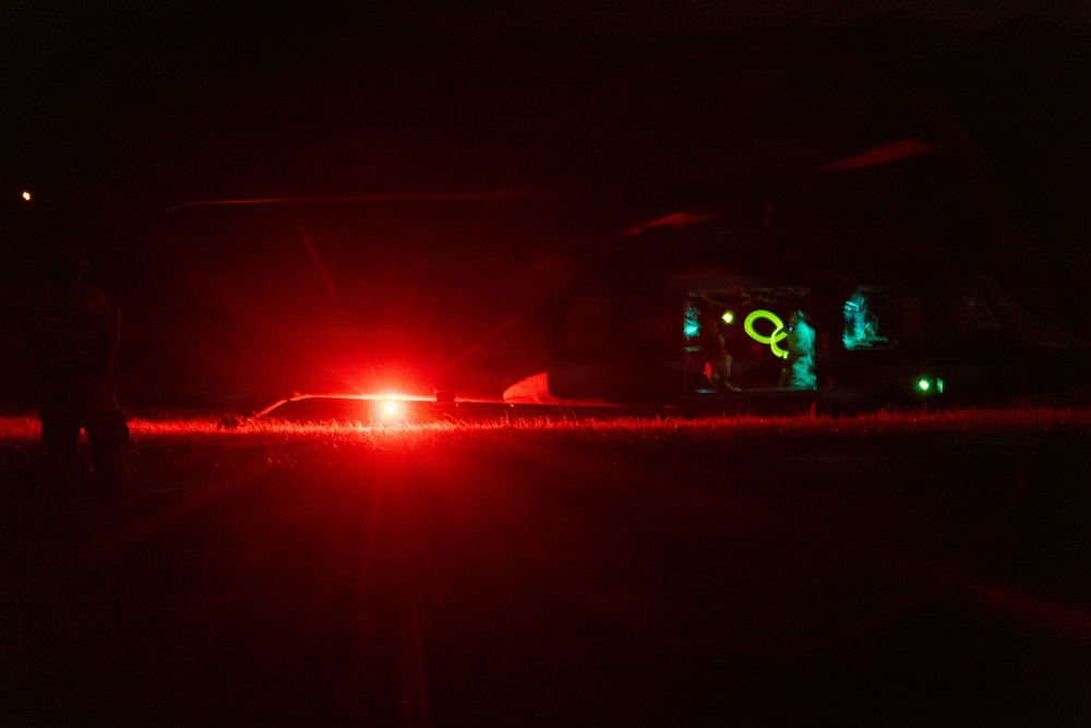 A nighttime outdoor scene featuring a large aircraft with an open rear cargo ramp illuminated by teal/cyan interior lighting. A bright red light source dominates the foreground, likely from a ground vehicle or beacon. Personnel are visible near the aircraft's open ramp area.