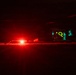 A nighttime outdoor scene featuring a large aircraft with an open rear cargo ramp illuminated by teal/cyan interior lighting. A bright red light source dominates the foreground, likely from a ground vehicle or beacon. Personnel are visible near the aircraft's open ramp area.