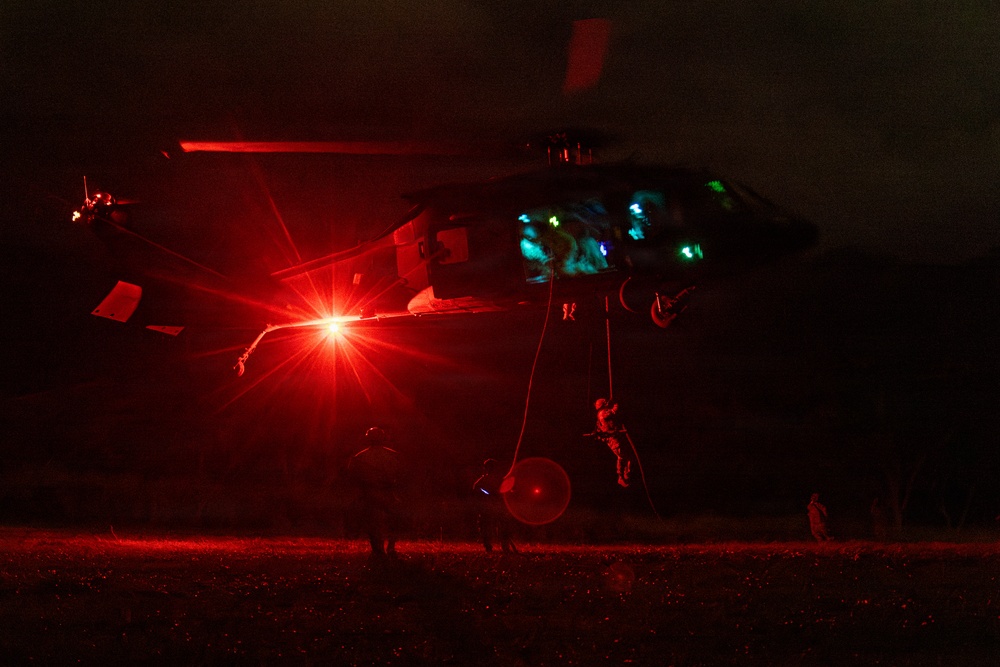 A military helicopter, consistent with a UH-60 Black Hawk, is hovering at low altitude during a nighttime operation. Personnel are rappelling or fast-roping down from the aircraft while ground personnel are visible below. The scene is dramatically illuminated by a bright red navigation/warning light on the helicopter, with teal/green cockpit lighting also visible.