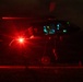 A military helicopter, consistent with a UH-60 Black Hawk, is hovering at low altitude during a nighttime operation. Personnel are rappelling or fast-roping down from the aircraft while ground personnel are visible below. The scene is dramatically illuminated by a bright red navigation/warning light on the helicopter, with teal/green cockpit lighting also visible.