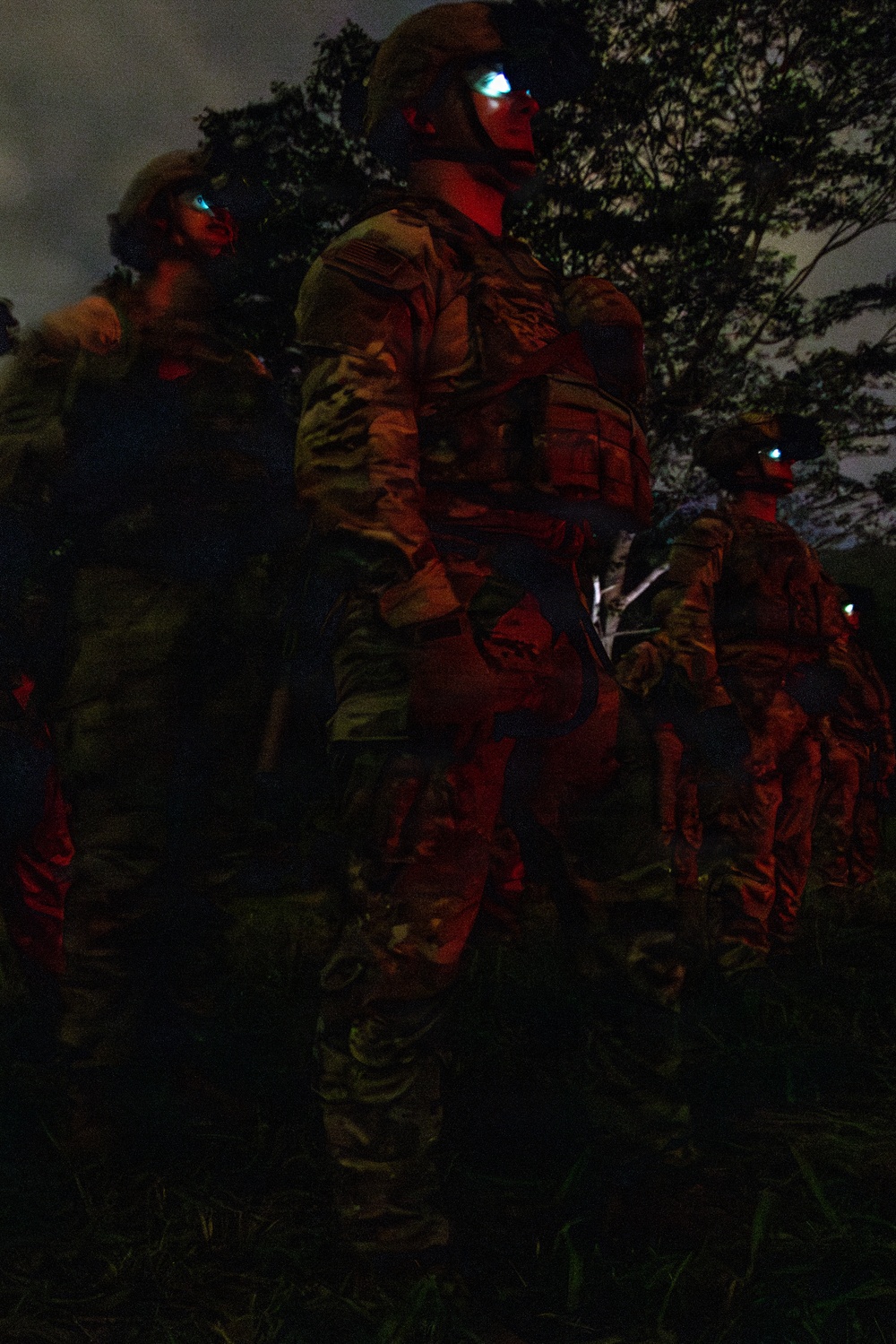 A group of military personnel in full combat gear are standing in formation during nighttime operations in a wooded or jungle environment. The soldiers are illuminated by red light, which is consistent with tactical night operations to preserve night vision. At least one soldier has a device mounted on their helmet emitting a blue-white light, likely night vision equipment.