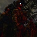 A group of military personnel in full combat gear are standing in formation during nighttime operations in a wooded or jungle environment. The soldiers are illuminated by red light, which is consistent with tactical night operations to preserve night vision. At least one soldier has a device mounted on their helmet emitting a blue-white light, likely night vision equipment.