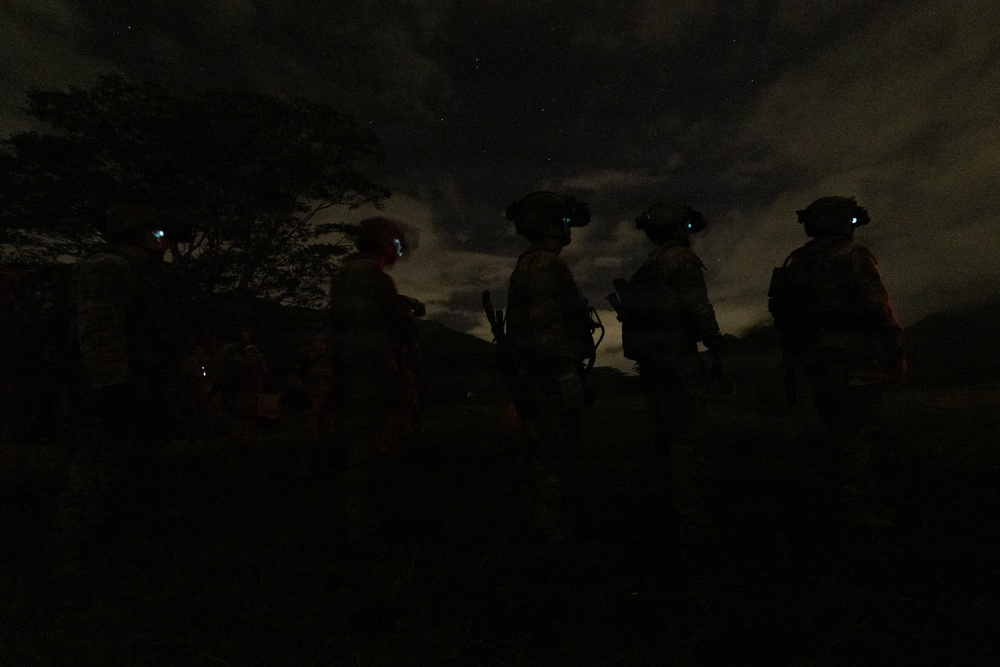 A nighttime military operation showing several soldiers in silhouette against a partially cloudy sky with visible stars. The personnel are equipped with helmets mounted with night vision devices (NVDs), tactical gear, and appear to be conducting a nighttime patrol or reconnaissance mission. The scene is extremely dark with figures only discernible as silhouettes.