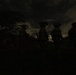 A nighttime military operation showing several soldiers in silhouette against a partially cloudy sky with visible stars. The personnel are equipped with helmets mounted with night vision devices (NVDs), tactical gear, and appear to be conducting a nighttime patrol or reconnaissance mission. The scene is extremely dark with figures only discernible as silhouettes.