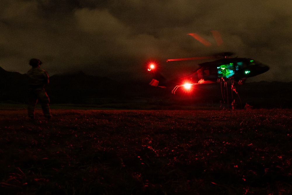A nighttime military helicopter operation captured in low-light conditions. A helicopter, likely a UH-60 Black Hawk, is on or near the ground with navigation lights illuminated, including a prominent red light and green cockpit lighting. At least one personnel figure is visible on the ground nearby.