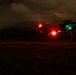 A nighttime military helicopter operation captured in low-light conditions. A helicopter, likely a UH-60 Black Hawk, is on or near the ground with navigation lights illuminated, including a prominent red light and green cockpit lighting. At least one personnel figure is visible on the ground nearby.