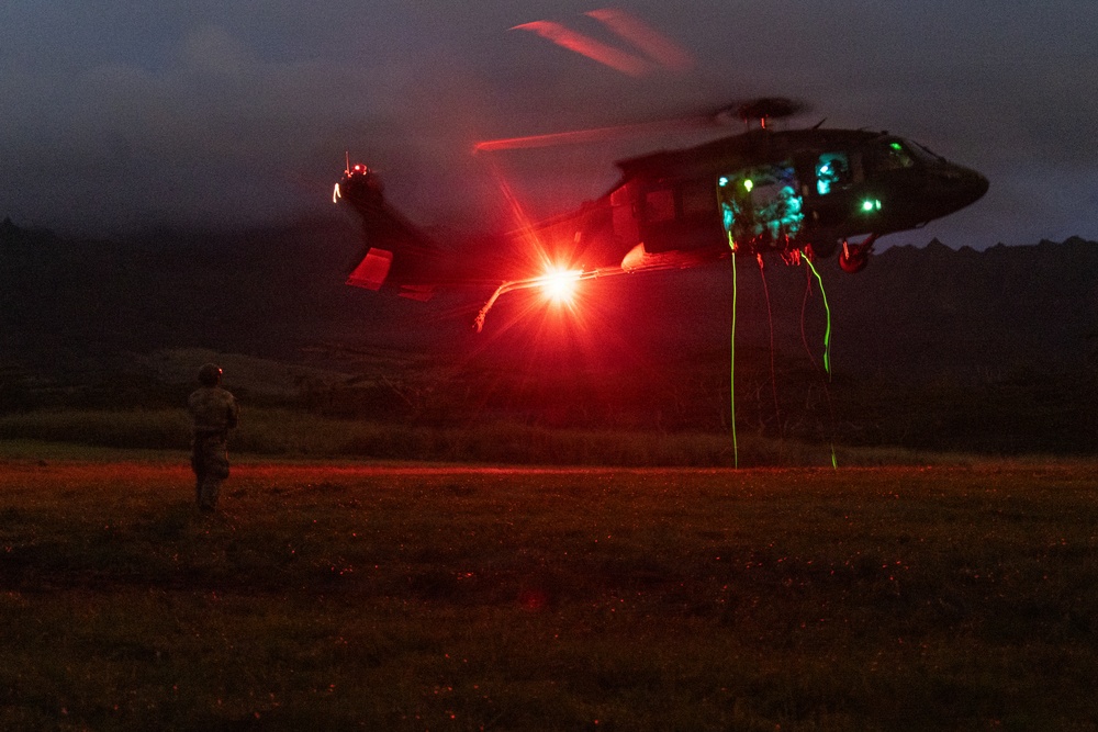A military helicopter, consistent with a UH-60 Black Hawk, hovers at low altitude during a nighttime operation over an open field. The scene is illuminated by a bright red light emanating from the aircraft, with green ropes or lines hanging from the fuselage suggesting a fast-rope or rappelling exercise. A single soldier in tactical gear is visible on the ground observing or preparing to interact with the aircraft.