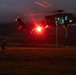 A military helicopter, consistent with a UH-60 Black Hawk, hovers at low altitude during a nighttime operation over an open field. The scene is illuminated by a bright red light emanating from the aircraft, with green ropes or lines hanging from the fuselage suggesting a fast-rope or rappelling exercise. A single soldier in tactical gear is visible on the ground observing or preparing to interact with the aircraft.