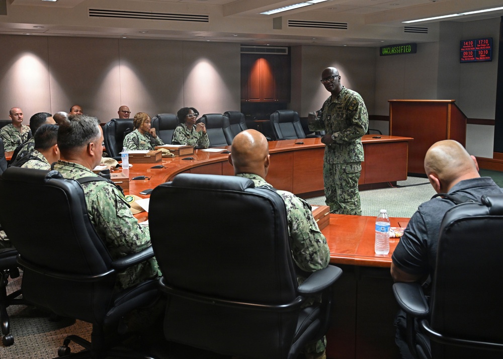 A military briefing or meeting is taking place in a formal conference room, with personnel wearing U.S. Navy camouflage uniforms (NWU Type III). One standing individual appears to be addressing the group seated around a large curved wooden conference table. A green 'UNCLASSIFIED' sign and digital clocks are visible on the back wall, indicating a secure but unclassified facility.