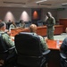 A military briefing or meeting is taking place in a formal conference room, with personnel wearing U.S. Navy camouflage uniforms (NWU Type III). One standing individual appears to be addressing the group seated around a large curved wooden conference table. A green 'UNCLASSIFIED' sign and digital clocks are visible on the back wall, indicating a secure but unclassified facility.