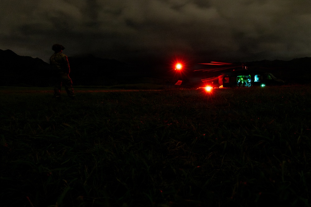 A nighttime military operation showing a soldier in camouflage standing near a helicopter on an open field. The helicopter displays red navigation/warning lights and green interior lighting, suggesting it is powered on and possibly preparing for takeoff or having just landed. The scene is set against a dark mountainous backdrop with overcast skies.