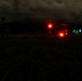 A nighttime military operation showing a soldier in camouflage standing near a helicopter on an open field. The helicopter displays red navigation/warning lights and green interior lighting, suggesting it is powered on and possibly preparing for takeoff or having just landed. The scene is set against a dark mountainous backdrop with overcast skies.