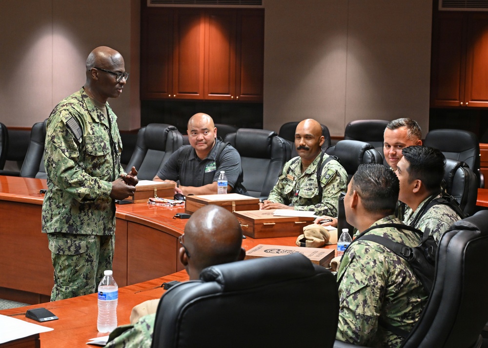 A U.S. Navy officer in NWU Type III (digital camouflage) uniform is addressing a small group of military personnel seated around a conference table in what appears to be a formal briefing room. The standing officer, whose name tape reads 'DAVIS,' appears to be conducting a meeting or briefing. Several attendees are in similar Navy working uniforms while at least one individual is in civilian attire.