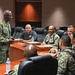 A U.S. Navy officer in NWU Type III (digital camouflage) uniform is addressing a small group of military personnel seated around a conference table in what appears to be a formal briefing room. The standing officer, whose name tape reads 'DAVIS,' appears to be conducting a meeting or briefing. Several attendees are in similar Navy working uniforms while at least one individual is in civilian attire.