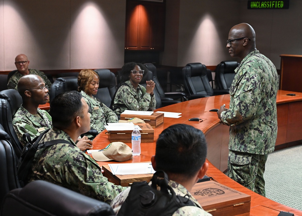 A military briefing or meeting is taking place in a conference room with personnel wearing U.S. Navy Working Uniform (NWU Type III) camouflage. A senior officer is standing and addressing a group of seated personnel around a large wooden conference table. A green LED display in the background reads 'UNCLASSIFIED', indicating a formal but non-classified meeting environment.