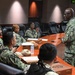 A military briefing or meeting is taking place in a conference room with personnel wearing U.S. Navy Working Uniform (NWU Type III) camouflage. A senior officer is standing and addressing a group of seated personnel around a large wooden conference table. A green LED display in the background reads 'UNCLASSIFIED', indicating a formal but non-classified meeting environment.