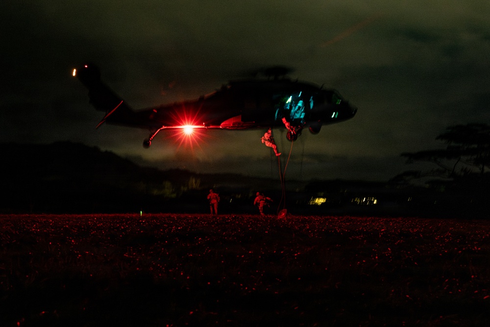 A nighttime military helicopter operation showing personnel fast-roping or rappelling from a hovering helicopter, illuminated by a bright red light source beneath the aircraft. The scene is captured in near-darkness with the ground bathed in red light, suggesting use of tactical lighting. Multiple personnel are visible at various stages of descent from the helicopter to the ground.