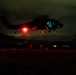 A nighttime military helicopter operation showing personnel fast-roping or rappelling from a hovering helicopter, illuminated by a bright red light source beneath the aircraft. The scene is captured in near-darkness with the ground bathed in red light, suggesting use of tactical lighting. Multiple personnel are visible at various stages of descent from the helicopter to the ground.
