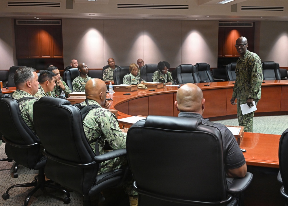 A military briefing or meeting is taking place in a formal conference room with a large curved wooden table. Multiple personnel wearing U.S. Navy NWU (Naval Working Uniform) Type III camouflage uniforms are seated around the table, while one individual stands and appears to be addressing the group. The setting suggests an official military command or administrative facility.