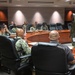 A military briefing or meeting is taking place in a formal conference room with a large curved wooden table. Multiple personnel wearing U.S. Navy NWU (Naval Working Uniform) Type III camouflage uniforms are seated around the table, while one individual stands and appears to be addressing the group. The setting suggests an official military command or administrative facility.