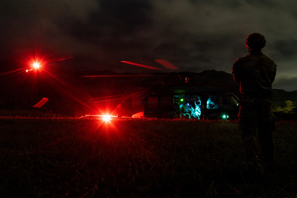A nighttime military helicopter operation captured in low-light conditions. A silhouetted soldier in tactical gear and helmet stands watch beside a large military helicopter, likely a UH-60 Black Hawk or similar rotary-wing aircraft, which is on the ground with its cabin illuminated. Bright red navigation/warning lights and interior blue-green lighting are the primary light sources in the scene.