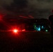 A nighttime military helicopter operation captured in low-light conditions. A silhouetted soldier in tactical gear and helmet stands watch beside a large military helicopter, likely a UH-60 Black Hawk or similar rotary-wing aircraft, which is on the ground with its cabin illuminated. Bright red navigation/warning lights and interior blue-green lighting are the primary light sources in the scene.