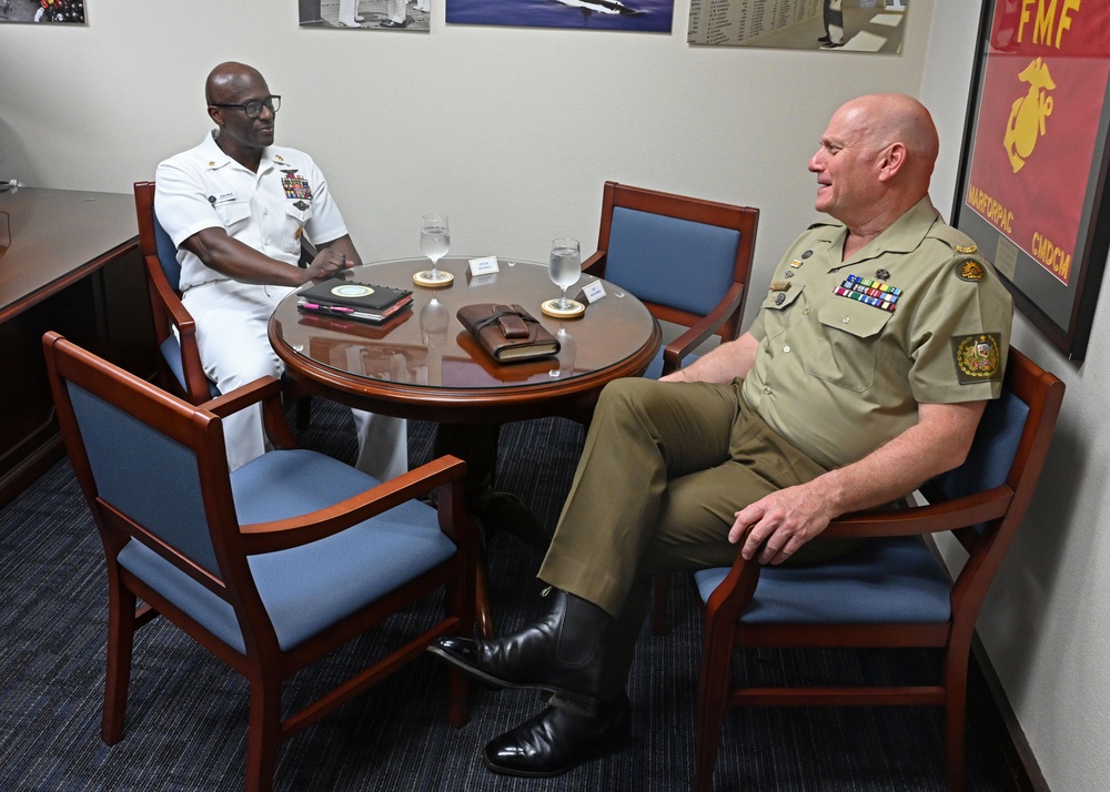 Two military officers are seated at a round conference table in what appears to be an official office setting. One officer is wearing a white U.S. Navy dress uniform with medals and ribbons, while the other is wearing an olive/khaki military uniform with a shoulder patch, consistent with Australian Defence Force dress. They appear to be engaged in a bilateral meeting or discussion.