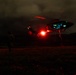 A nighttime scene showing a military helicopter in low hover or landing phase over an open field. A single person stands on the ground nearby, illuminated by the helicopter's bright red navigation/landing light. The scene is extremely dark with the aircraft and figure only visible due to the aircraft's navigation lights.