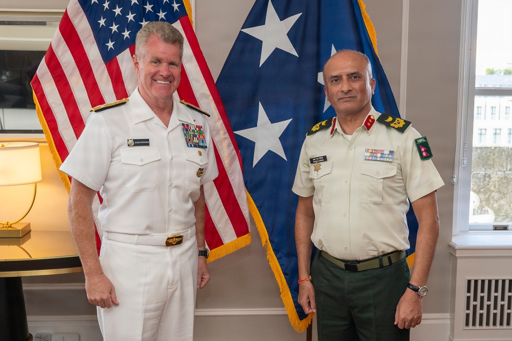 Two military officers in formal dress uniforms pose for a photo in what appears to be an official office setting. The officer on the left wears a U.S. Navy white dress uniform with a name tag reading 'SAM PAPARO' and multiple ribbons, while the officer on the right wears a light khaki uniform with green trousers bearing a 'NEPAL' patch. They are standing in front of a U.S. flag and a blue flag with white stars.
