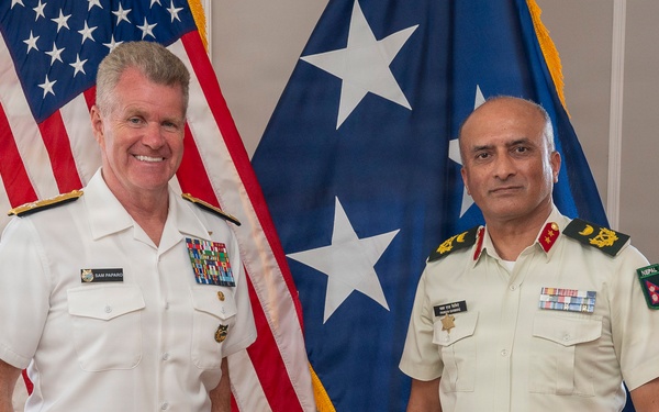 Two military officers in formal dress uniforms pose for a photo in what appears to be an official office setting. The officer on the left wears a U.S. Navy white dress uniform with a name tag reading 'SAM PAPARO' and multiple ribbons, while the officer on the right wears a light khaki uniform with green trousers bearing a 'NEPAL' patch. They are standing in front of a U.S. flag and a blue flag with white stars.