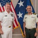 Two military officers in formal dress uniforms pose for a photo in what appears to be an official office setting. The officer on the left wears a U.S. Navy white dress uniform with a name tag reading 'SAM PAPARO' and multiple ribbons, while the officer on the right wears a light khaki uniform with green trousers bearing a 'NEPAL' patch. They are standing in front of a U.S. flag and a blue flag with white stars.