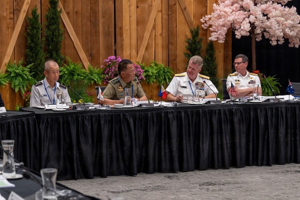 A formal multinational military conference is taking place at a long draped table. Four uniformed military officers from different nations are seated together, with small national flags visible on the table indicating international representation. The setting appears to be a decorated conference venue with floral and wooden barn-style backdrop elements.