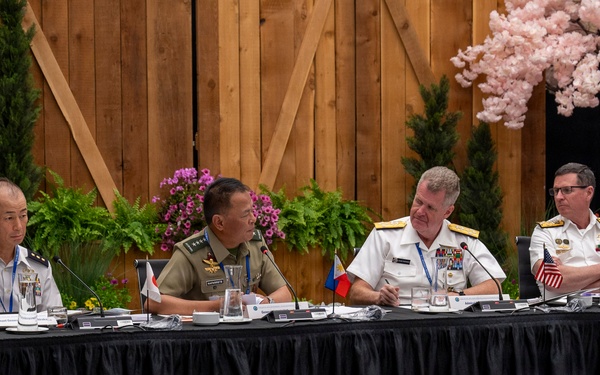 A formal multinational military conference is taking place at a long draped table. Four uniformed military officers from different nations are seated together, with small national flags visible on the table indicating international representation. The setting appears to be a decorated conference venue with floral and wooden barn-style backdrop elements.