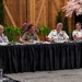 A formal multinational military conference is taking place at a long draped table. Four uniformed military officers from different nations are seated together, with small national flags visible on the table indicating international representation. The setting appears to be a decorated conference venue with floral and wooden barn-style backdrop elements.