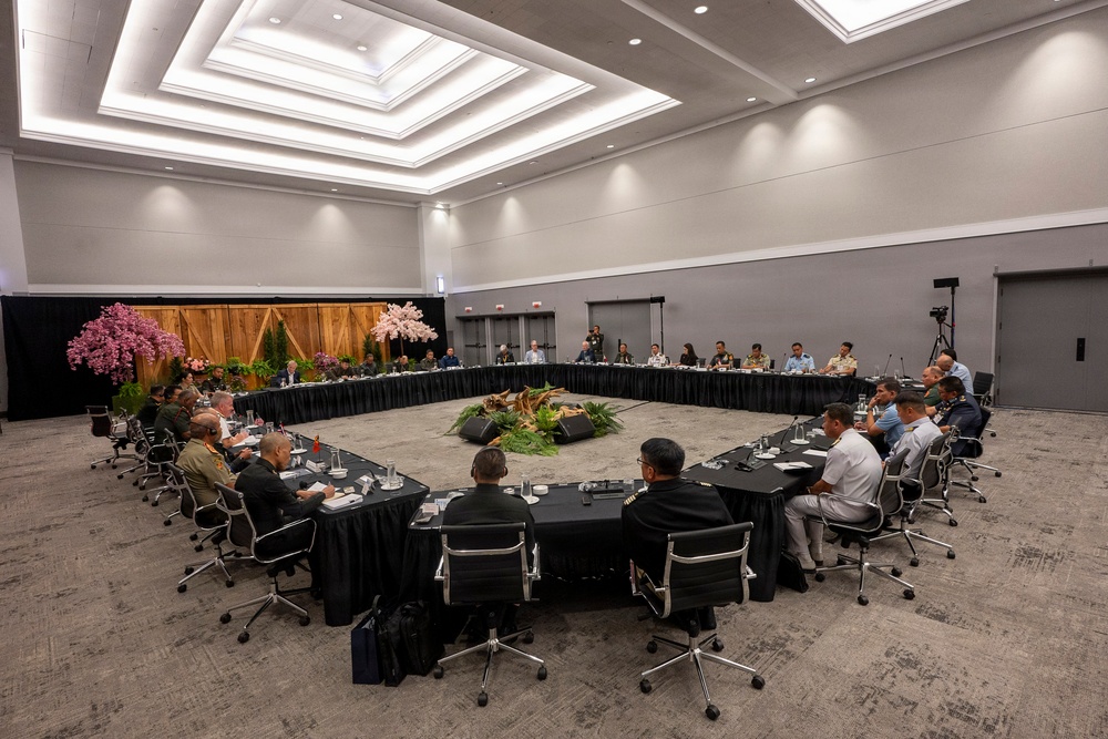 A large formal multilateral meeting or conference is taking place in a modern conference hall, with participants seated around a large square arrangement of tables with black skirting. The attendees appear to include military personnel in various uniforms and civilians, suggesting a high-level defense or diplomatic forum. The room is professionally decorated with floral arrangements and tropical plants in the center of the table configuration.